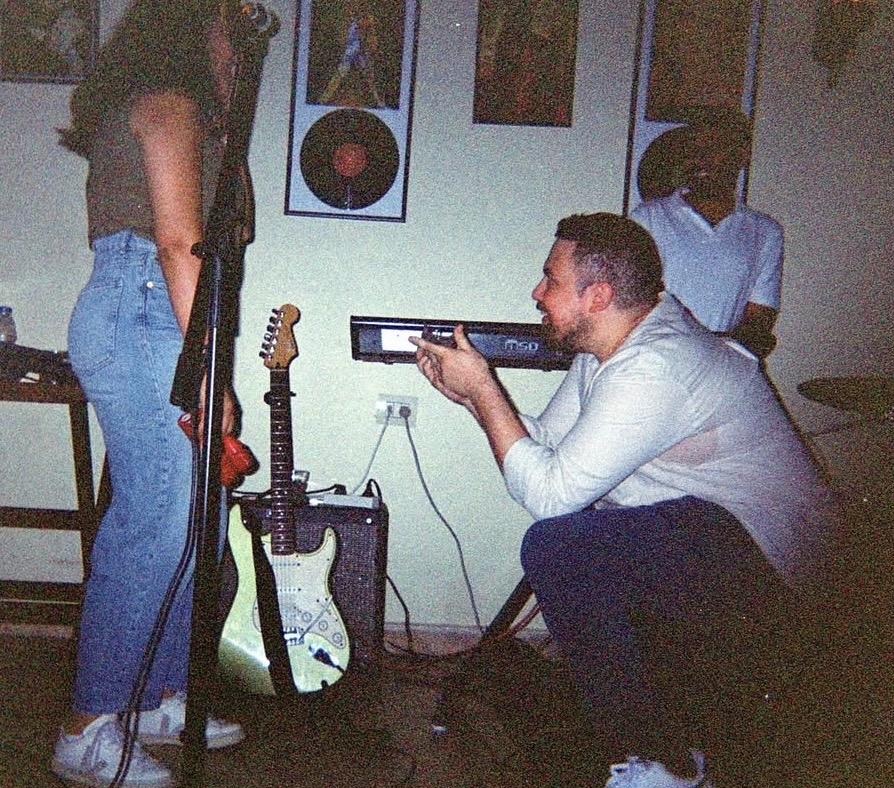 Roger crouched by an amplifier during soundcheck, guitar resting on its stand.